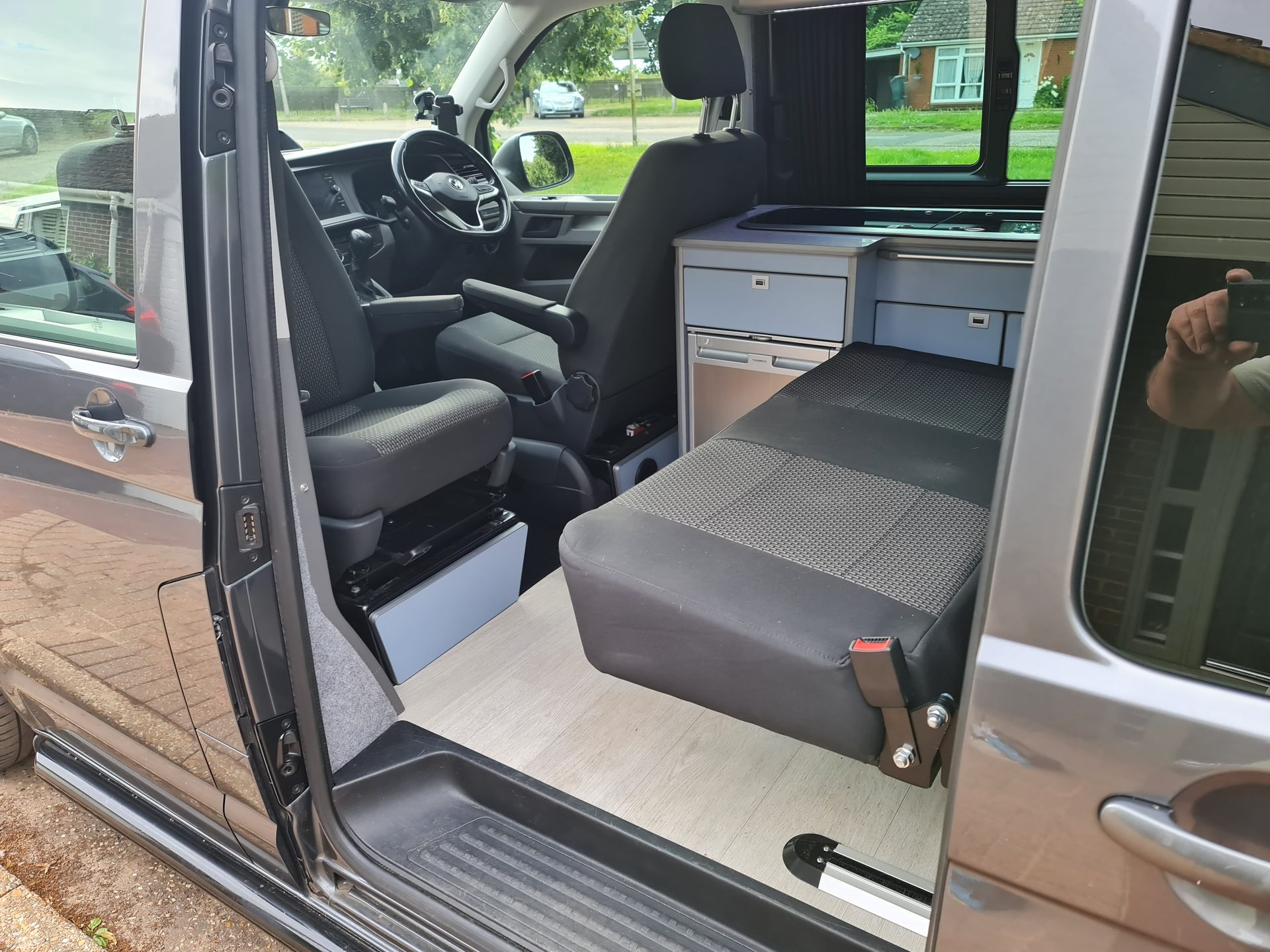 Open sliding door of a grey VW Transporter camper showing fold-out bed and pale blue kitchen units