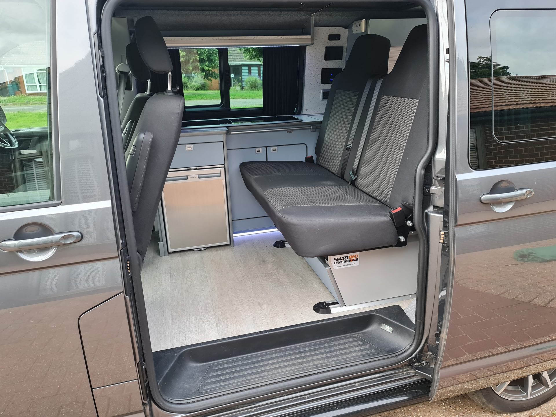 Open sliding door of a grey VW Transporter campervan showing rear bench seat, fridge and grey kitchen units
